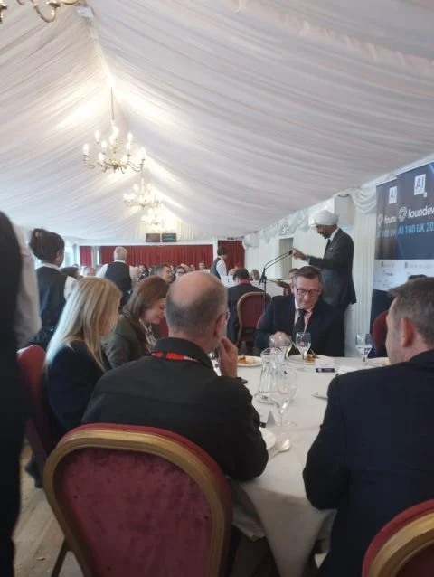 Guests seated at round tables in a marquee as Lord Kulveer Ranger addresses them beside a Foundever AI 100 UK banner